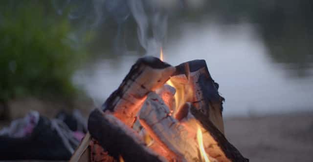 Some burning logs have been placed leaning together in a conical shape and are burning a deep red glow with a small wisp of smoke rising up and a lake in the background. If you have a burning question here are the contact details for Meet the Locals