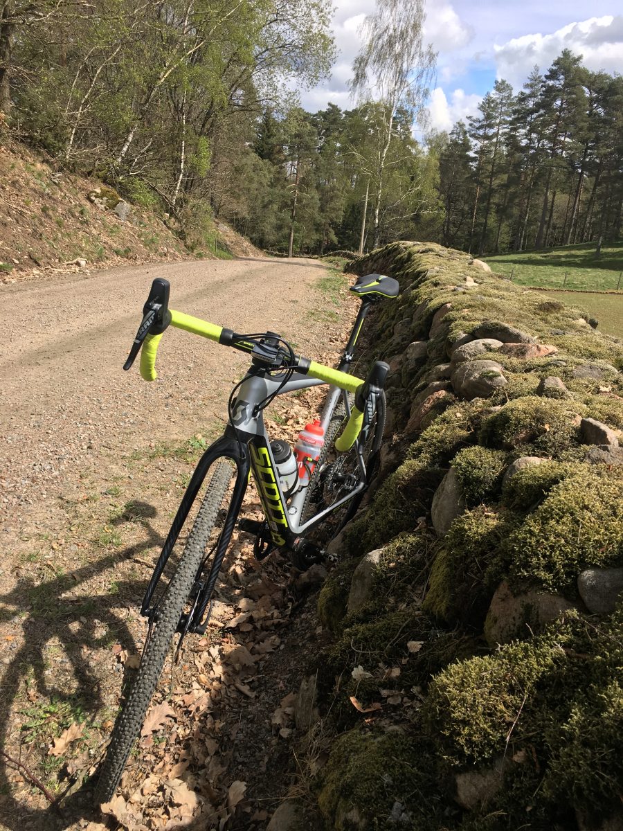 A riderless mountain bike leans against a mossy stone wall on a gravel road in the sunshine