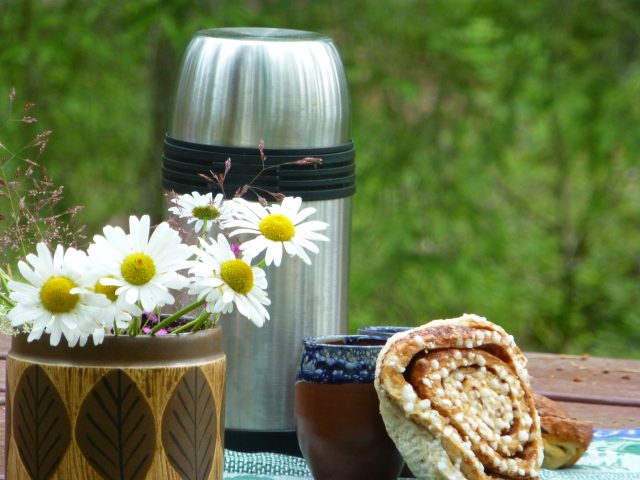 A stainless steel thermos flask stands on a picnic table beside a handmade mug and a cinnamon bun. There is also a brown vase from the 1970s with some bright white and yellow daisies. It must be time for a fika while hiking with a host from Meet the Locals