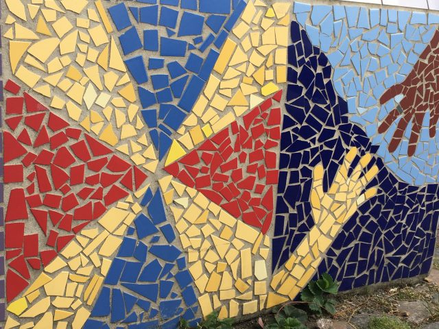 A mosaic mural made with broken up tiles shows a geometric pattern in blue, red, and yellow and also two hands reaching for each other. Some of the art that you might find on a tour with a host from Meet the Locals